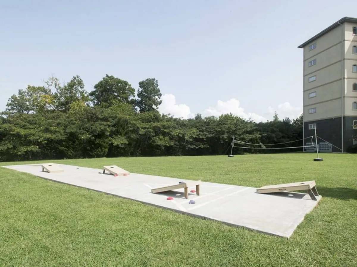 A playful exterior shot showing cornhole boards on a large lawn, adjacent to a building structure. The open green space provides plenty of room for games and community activities, capturing the spirit of outdoor fun and togetherness.