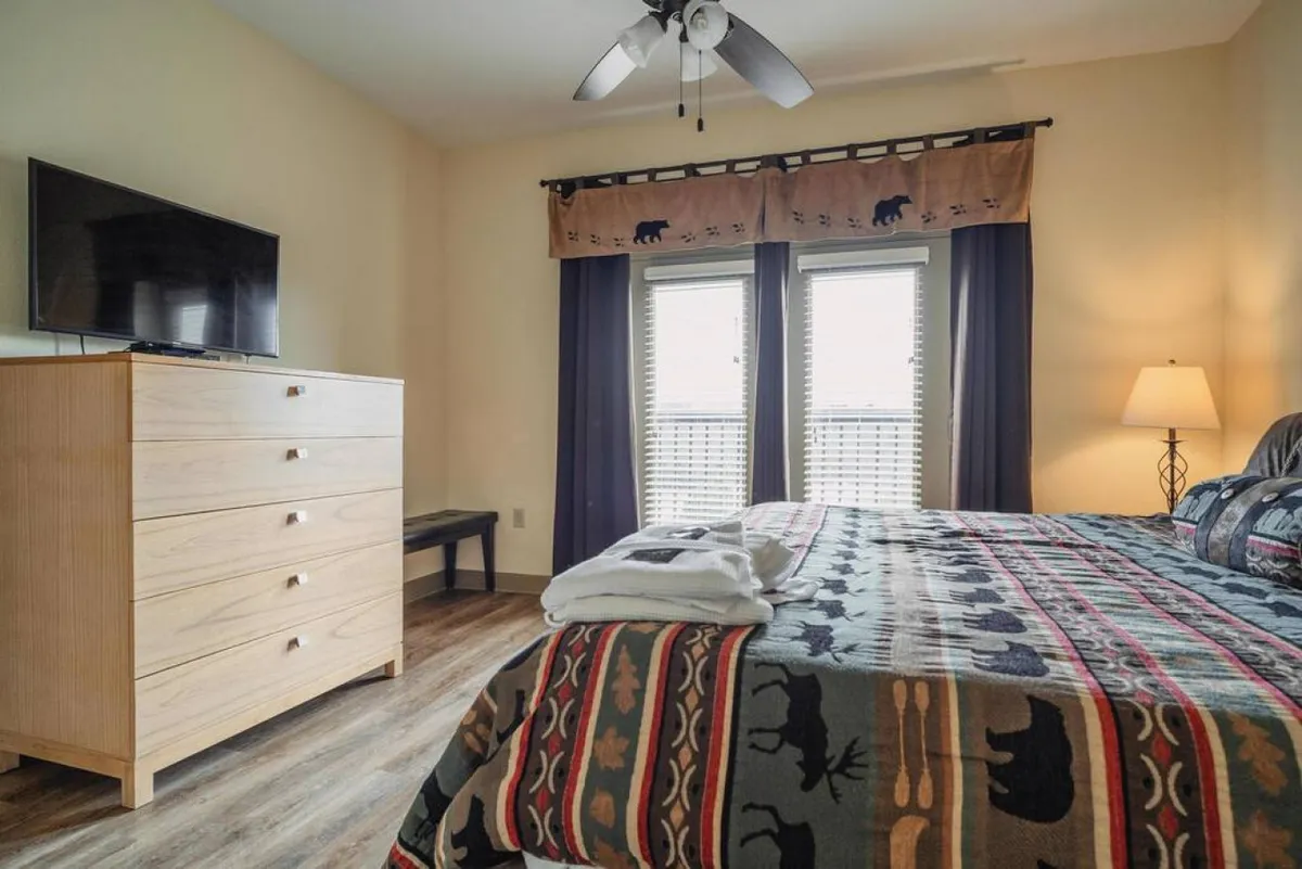 A warmly lit bedroom featuring a bed with a patterned blanket, a dresser, windows with curtains letting in natural light, and a wall-mounted TV. The atmosphere is cozy and inviting, ideal for relaxing after a day of activities.