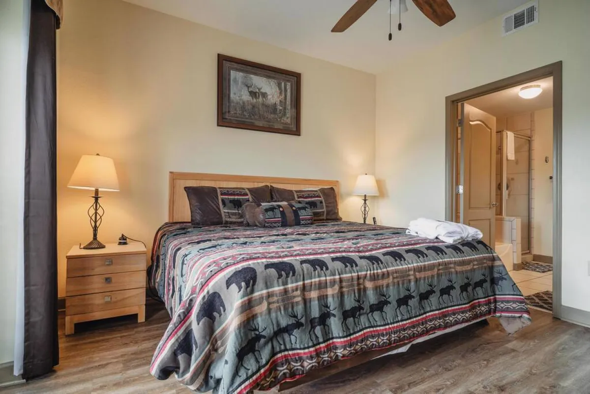 A tranquil and inviting bedroom with a queen bed dressed in patterned bedding, a nightstand topped with a lamp, and framed artwork on the wall. Soft, natural light enhances the serene mood, perfect for a peaceful night’s rest.