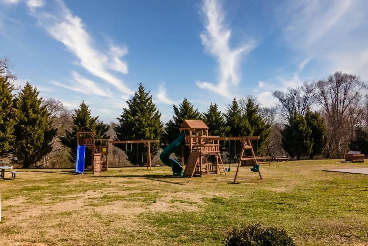 A cheerful exterior playground scene with colorful play equipment, swings, a slide, green grass, and shaded areas under mature trees. Children are actively playing while parents and families gather nearby, evoking a scene full of energy and joy.