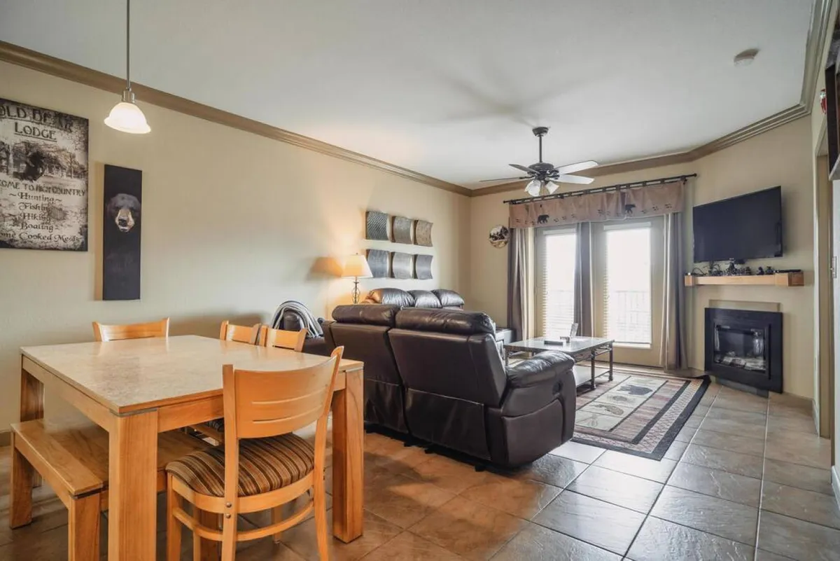 A warm, inviting living and dining area with a fireplace, large windows that let in natural light, a leather couch, and a dining table. The windows likely provide views of the surrounding landscape and shifting seasonal beauty, while the fireplace evokes cozy winter gatherings.