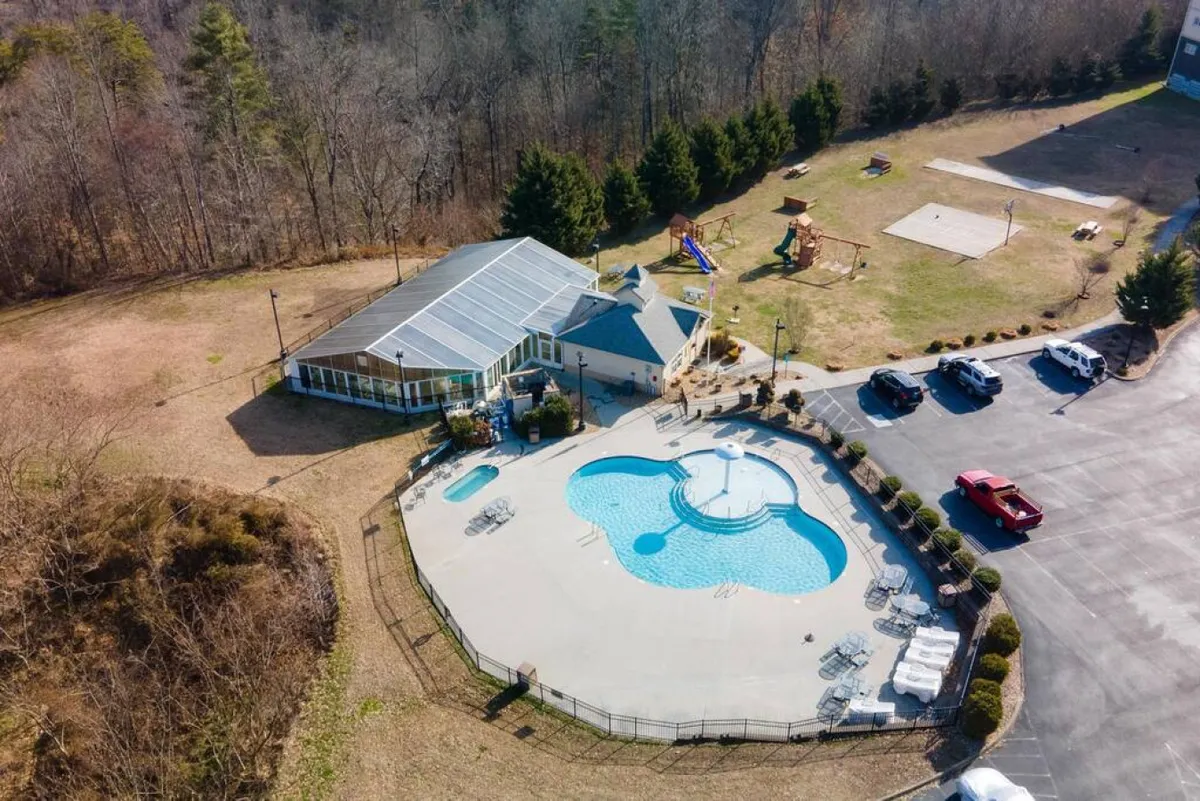 A lively exterior scene featuring an outdoor swimming pool surrounded by lounge chairs, a nearby recreation area with playground equipment, and a clubhouse in the background. Families and children are seen enjoying the pool, playing on the playground, and gathering outdoors, all under bright, sunny skies.