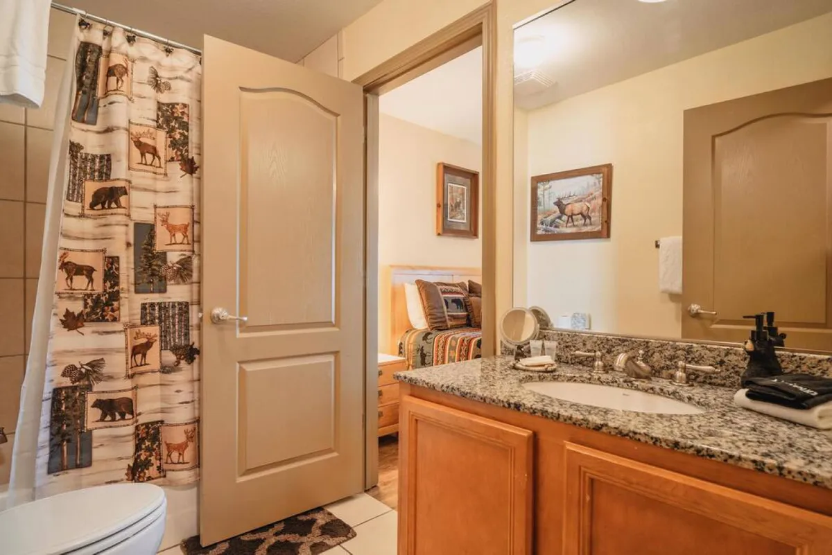 A cozy bathroom with a granite countertop, shower with a wildlife-themed curtain, wooden cabinetry, and a mirror reflecting a glimpse of a bedroom. The room feels inviting and practical, perfect for family routines.