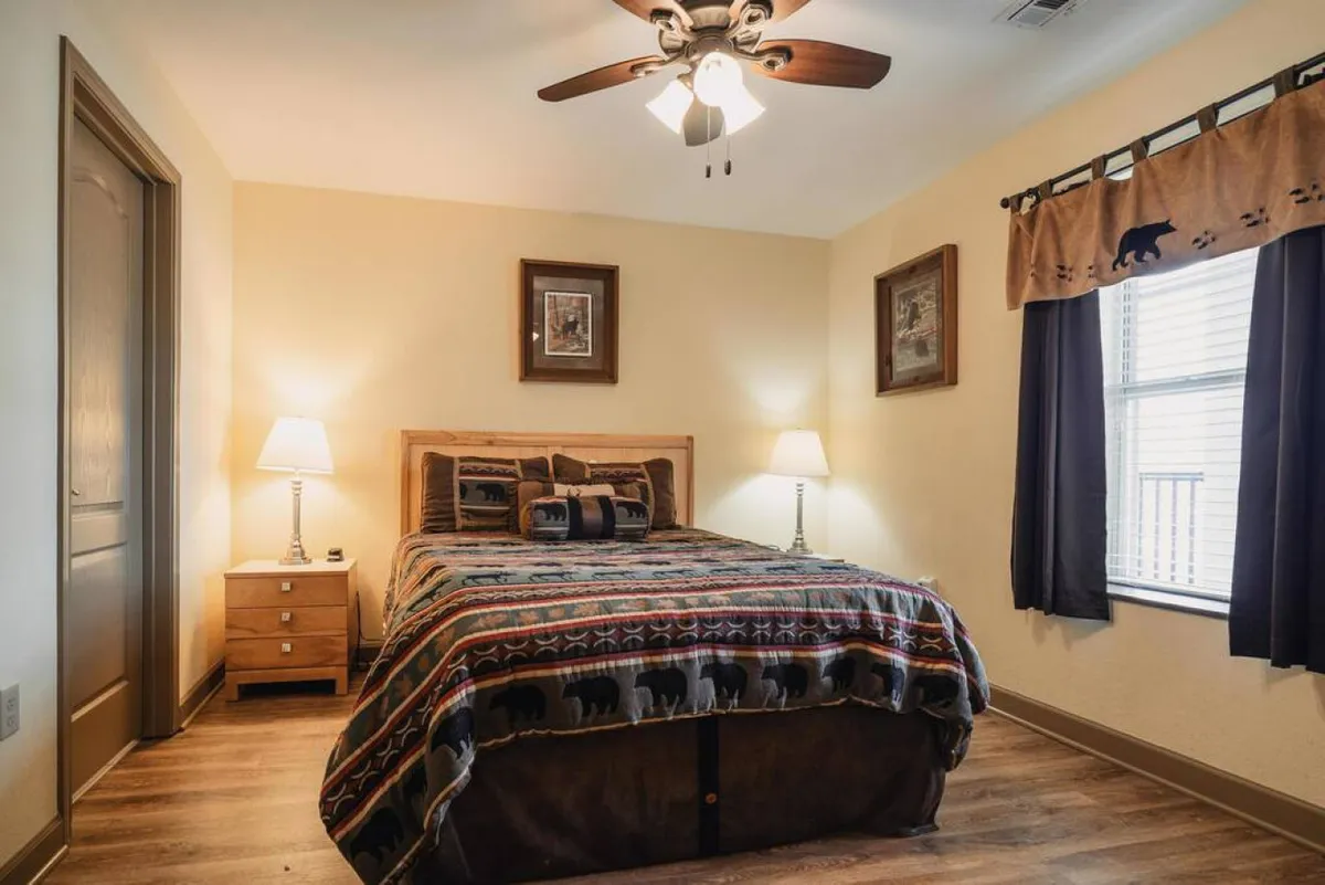 The image showcases a beautifully appointed bedroom with a plush, patterned comforter on a king-size bed, flanked by elegant nightstands and softly glowing lamps. Framed artwork adds a touch of sophistication, while a large window with flowing curtains invites in natural light and offers glimpses of the tranquil Smoky Mountain surroundings.